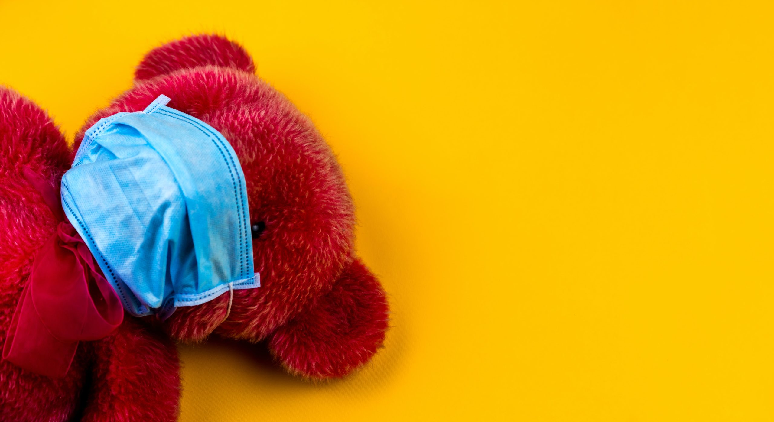 Einsamer roter Teddybär auf gelbem Hintergrund mit medizinischer Atemschutzmaske auf.