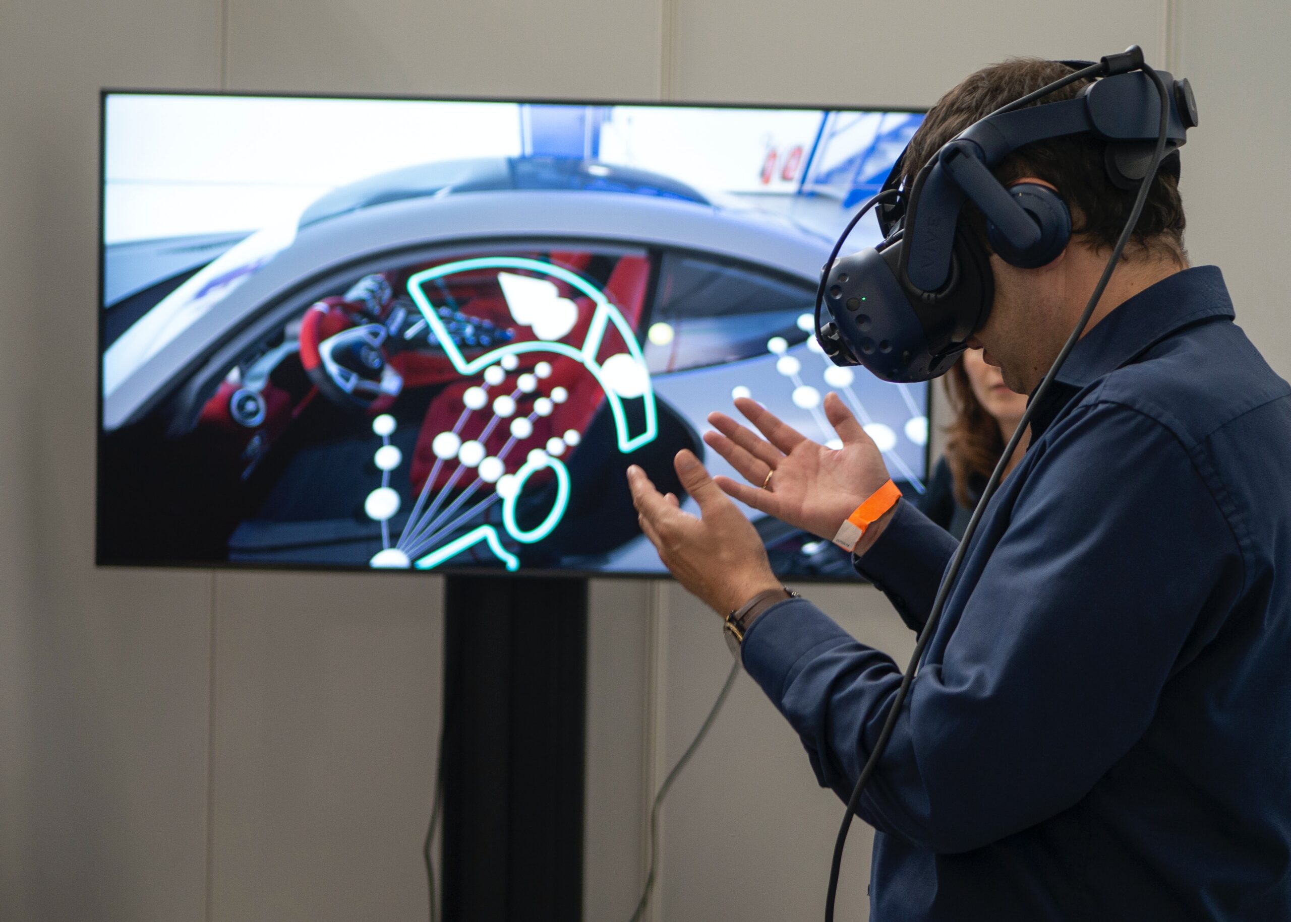 Ein Mann mit einer aufgezogenen VR Brille steht vor einem Monitor, auf dem das Bild aus seiner VR Brille abgebildet wird