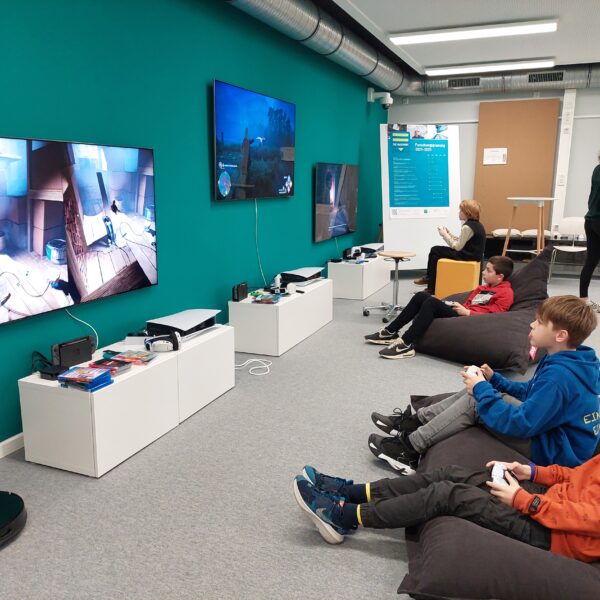 Eine Reihe Kinder sitzt am rechten Bildrand in Sitzsätzen und spielt an Fernsehern an der Wand auf der linken Seite Videospiele